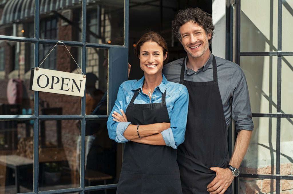 Two smiling people wearing black aprons stand outside a shop with an OPEN sign hanging on the glass door, suggesting they are owners or employees welcoming customers.