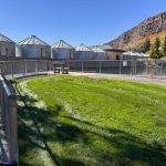Buhl ID 25 A fenced grassy area with a bench, surrounded by metal silos and rocky hills under a clear blue sky.