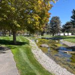 Buhl ID 24 A small stream with rocks runs through a grassy park, bordered by trees with green and yellow leaves under a clear blue sky. A paved walkway curves along the left side of the image.