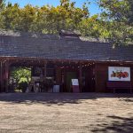 Buhl ID 10 A rustic wooden fruit stand with a shingled roof sits under leafy trees. A sign reads Kelleys Fruit, Jams, Jelly, Gifts with fruit illustrations. An Open sign stands by the entrance.