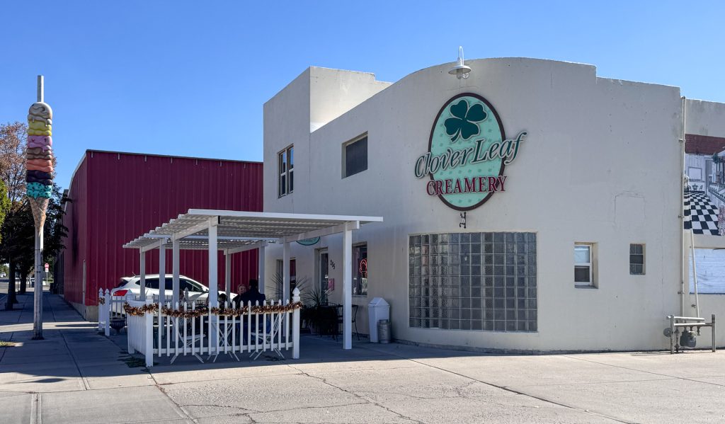 A white building with a large “CloverLeaf Creamery” sign, outdoor seating under a pergola, and a red building in the background, on a sunny day. A tall ice cream cone sculpture stands near the street.