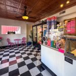 Buhl ID 12 A retro-style ice cream shop with black-and-white checkered floors, a counter with ice cream tubs, colorful “MILK” letters, a menu on the wall, and pink chairs around a small table by a window.