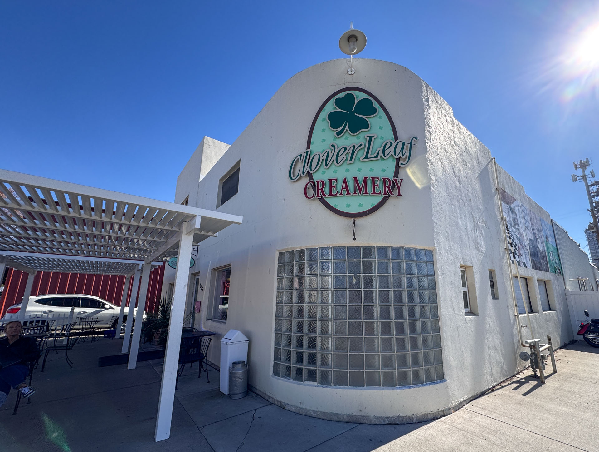 A white building with a round glass block window displays a large oval sign reading CloverLeaf Creamery with a green clover logo. Patio seating and a white pergola are visible in front, under a clear blue sky.