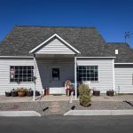 Buhl ID 22 Small white building with gray shingles, a central front door, potted plants, a mailbox, and traffic cones on the right side, under a clear blue sky. The area around the building is paved with gravel and concrete.