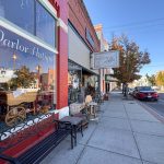 Buhl ID 14 A sunny street with antique shops, including “Parlor Antiques,” displaying vintage items outside. A red car is parked along the sidewalk, and trees with autumn leaves line the street. The sky is clear and blue.