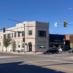 Buhl ID 16 A sunny street corner with a beige two-story building, several cars parked and driving, traffic lights, and signs reading “Broadway Ave.” and “N.” Smaller storefronts line the street under a clear blue sky.