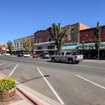Buhl ID 18 A small-town main street with parked cars, brick buildings, storefronts, and a green awning reading “FURNITURE & APPLIANCES.” A lamp post, flower planter, and clear blue sky are visible.