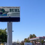 Buhl ID 21 A sign reads Trout Capital of the World, Buhl Chamber of Commerce with a trout illustration. Trees and a gas station with a Route 66 sign are visible under a clear blue sky.