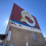 Buhl ID 20 A large mural of a fish jumping through a ring over water and sand is painted on the side of a brick building. A Thrift Store sign hangs above a wheelchair-accessible entrance. The sky is clear and sunny.