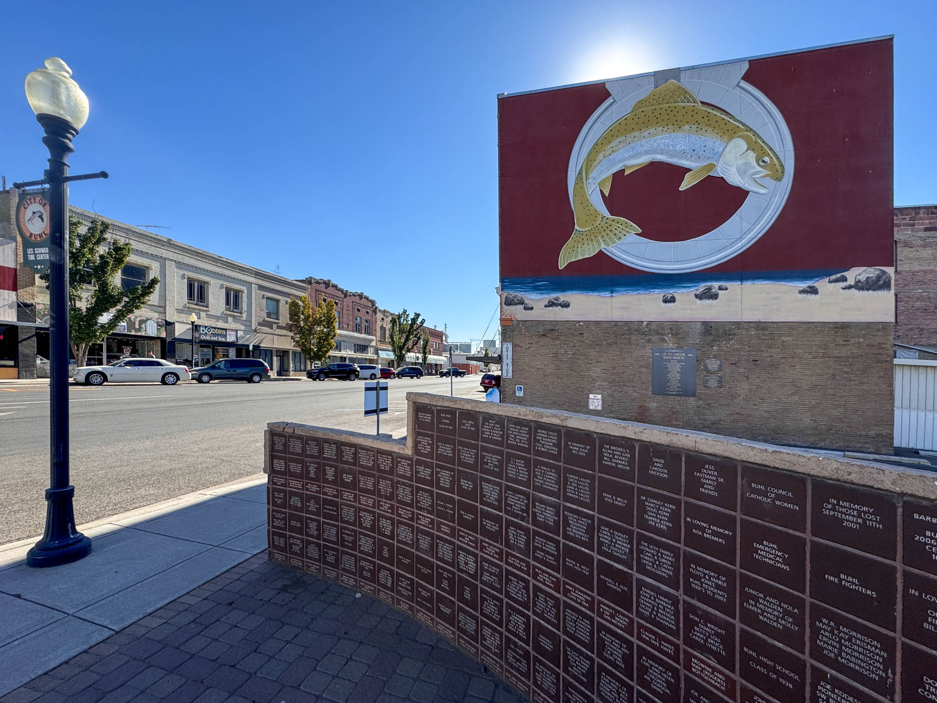A street scene with buildings and cars, featuring a large mural of a trout above a wall of engraved plaques. A vintage-style streetlamp stands in the foreground on the left. The sky is clear and sunny.