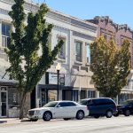 Buhl ID 19 A row of historic brick buildings with shops on the ground floor lines a sunny street. Several cars are parked along the curb, and leafy trees partially shade the storefronts.