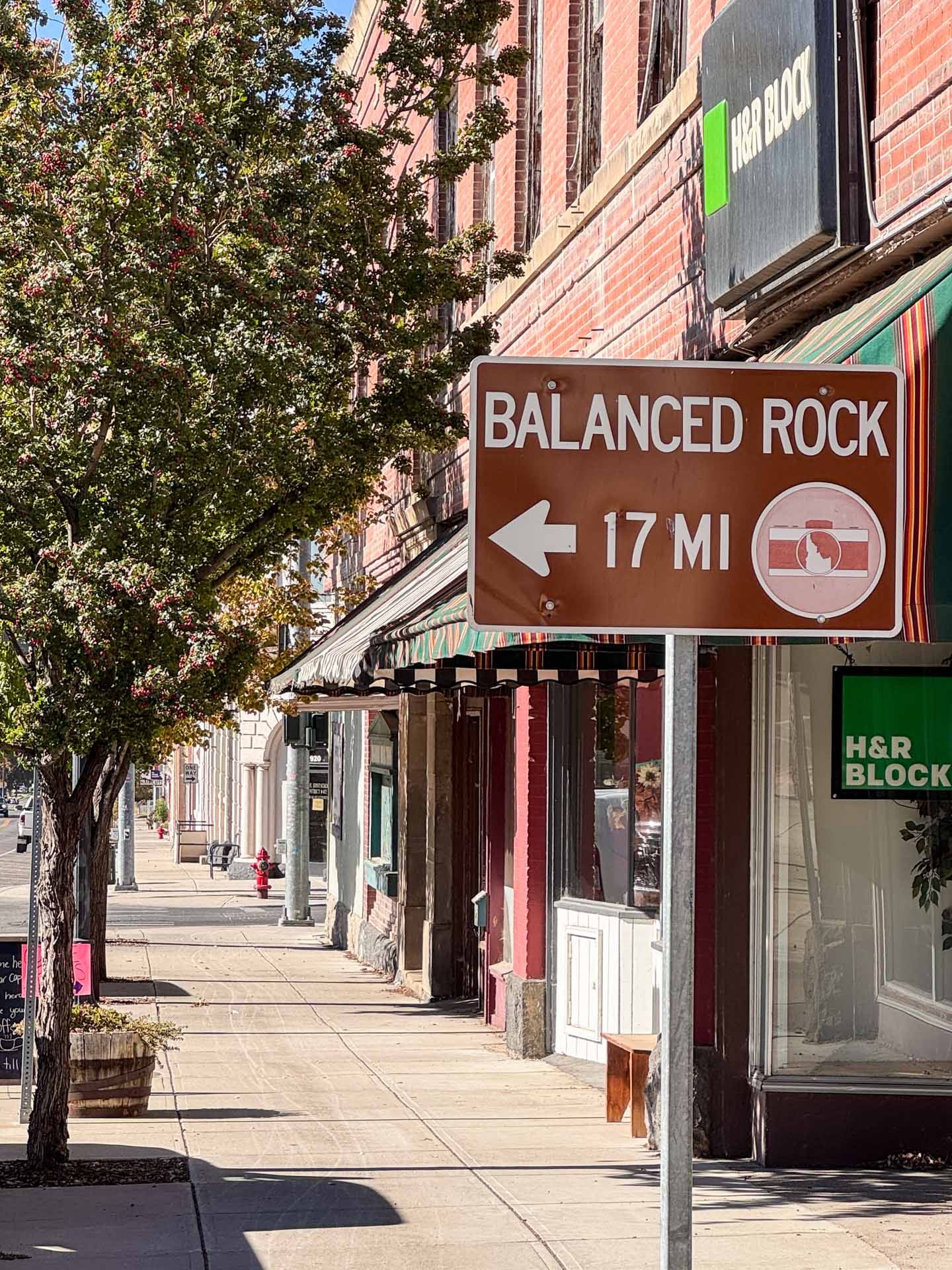 A brown road sign on a sidewalk points left, reading Balanced Rock 17 mi. Storefronts, including H&R Block, and trees line the sunny street in a small downtown area.