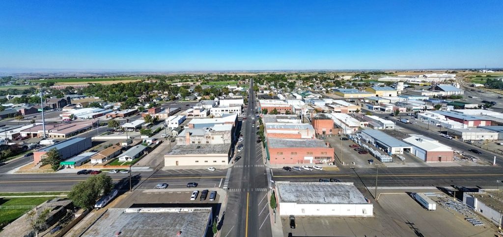 Aerial view of a small town with low-rise buildings, streets, parked cars, and green fields in the background under a clear blue sky.