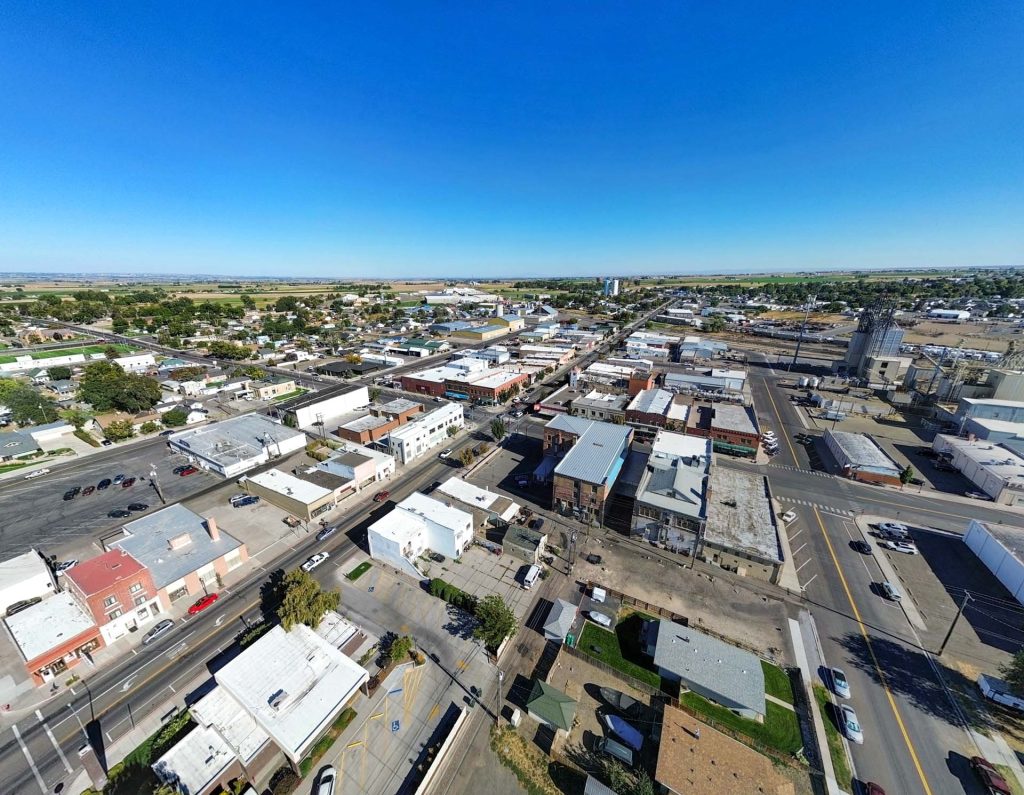 Aerial view of a small town with grid-like streets, low-rise buildings, and parked cars under a clear blue sky, surrounded by flat rural farmland.