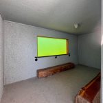 Buhl ID 27 A small, empty room with textured white walls, a wooden bench, and a wide rectangular yellow-tinted window. The room has a simple light fixture on the ceiling and a bare concrete floor.