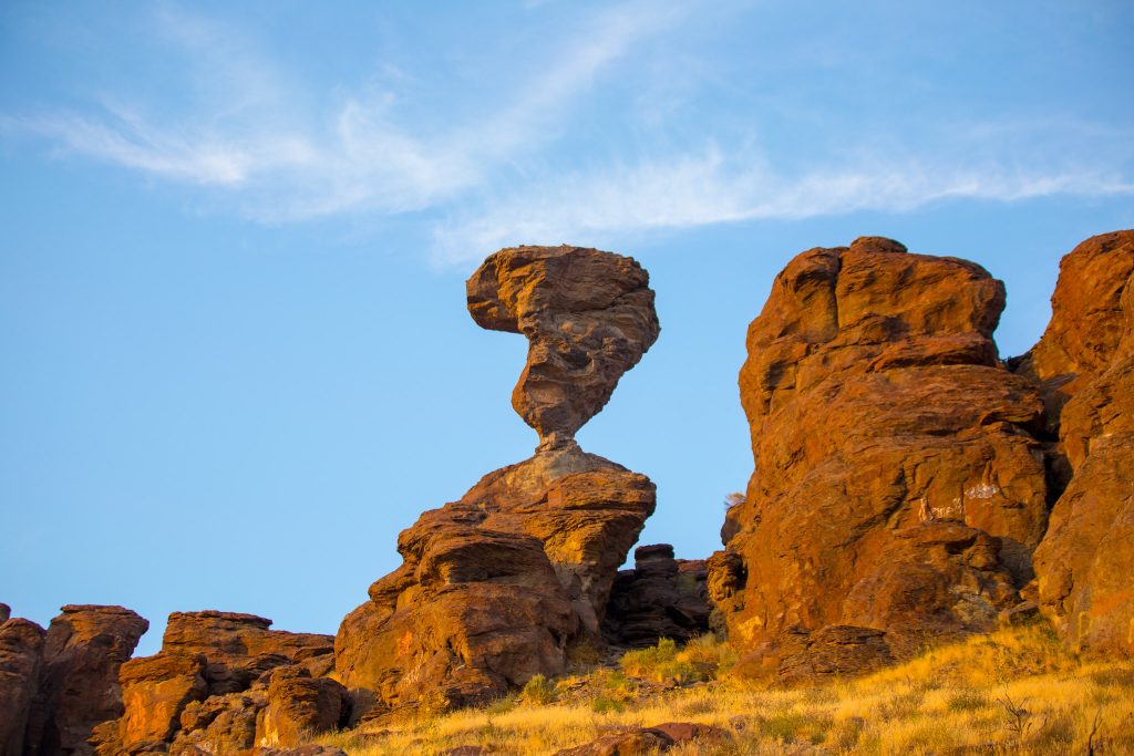 A tall, narrow rock formation, known as Buhl Rock, balances on a rocky ledge against a clear blue sky, surrounded by reddish-brown cliffs and dry grass.